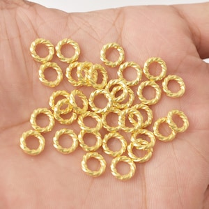 May include: A collection of small, gold-colored, twisted-design jump rings. These jewelry findings are scattered across a light-colored surface. The rings have a textured, rope-like appearance, and are likely used for jewelry making or crafting.