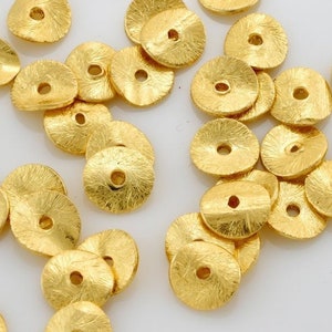 May include: A selection of small, gold-coloured, textured metal beads. Each bead is disc-shaped with a central hole, and the surface has a hammered or textured appearance. The beads are scattered on a white surface.