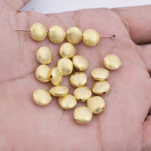 May include: A collection of gold-colored round beads with a textured surface. The beads are scattered on a hand.