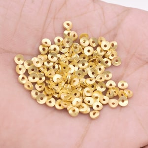 Puede incluir: Un montón de cuentas de metal de color dorado en forma de anillo. Las cuentas son pequeñas y tienen una superficie ligeramente texturizada.