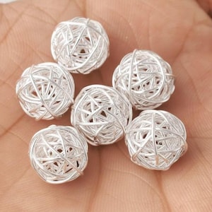 Puede incluir: Seis cuentas esféricas plateadas con un diseño abierto y tejido. Las cuentas están hechas de finos hilos de metal entrelazados, creando una apariencia delicada y aireada. Las cuentas miden aproximadamente 2 cm de diámetro, adecuadas para la fabricación de joyas o proyectos de artesanía.