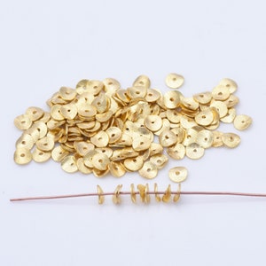May include: A pile of small, gold-coloured, disc-shaped beads with a textured surface and a central hole. Some beads are strung on a thin, copper-coloured wire, demonstrating their use in jewellery making. The beads are scattered on a white surface.