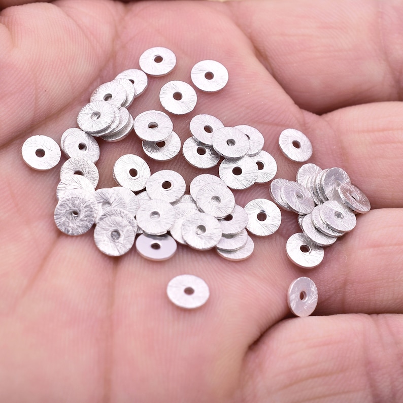 May include: A pile of silver metal round beads with a textured surface. The beads are scattered on a hand.