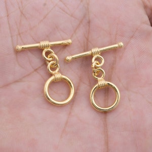 May include: Two gold-toned metal toggle clasps with round rings. Each clasp has a straight bar with a rounded end and a small ring attached to the bar.