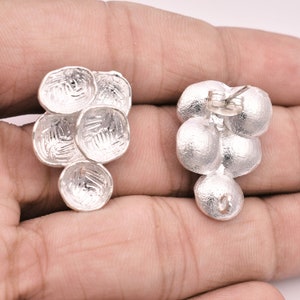 May include: A pair of silver earrings with a cluster of round, textured discs. The earrings have a post and butterfly back closure.
