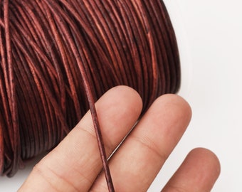 May include: A spool of dark brown leather cord. The cord is thin and smooth, and it is wound tightly around the spool. A hand is holding a piece of the cord, showing its texture.