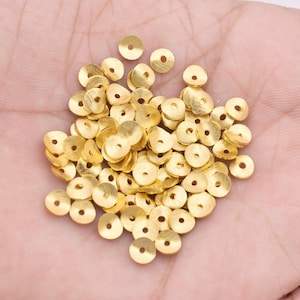 May include: A pile of gold-coloured metal beads, each with a small hole in the centre. The beads are round and flat, with a slightly textured surface.