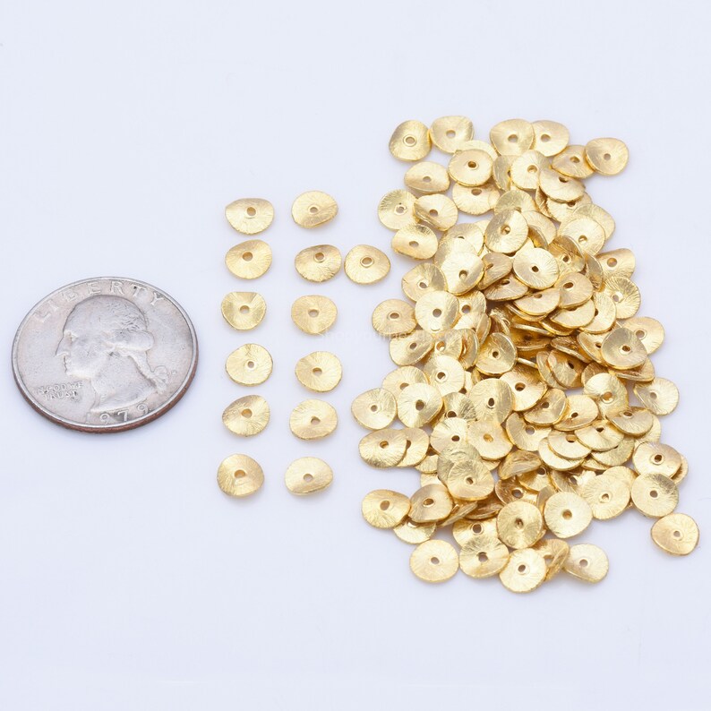 May include: A scattering of small, gold-coloured, disc-shaped beads with a central hole, on a white surface. A U.S. quarter coin is next to the beads for size comparison. These beads are likely for jewellery making or other craft projects.