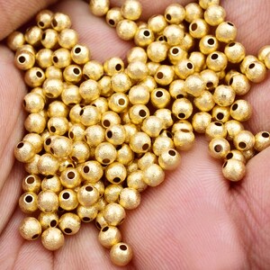 May include: A large pile of gold-colored metal beads with a textured surface. The beads are round and have a hole in the center.