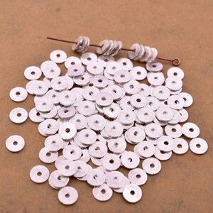May include: A large pile of silver-colored metal spacer beads. The beads are round with a hole in the center. The beads are scattered on a brown surface.