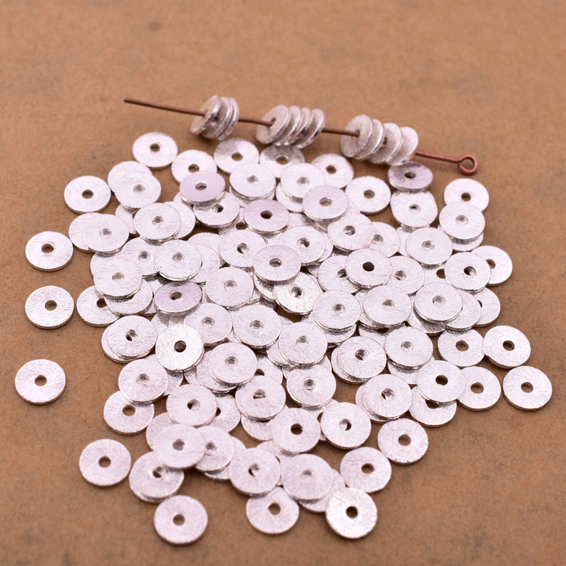 May include: A large pile of silver-colored metal spacer beads. The beads are round with a hole in the center. The beads are scattered on a brown surface.