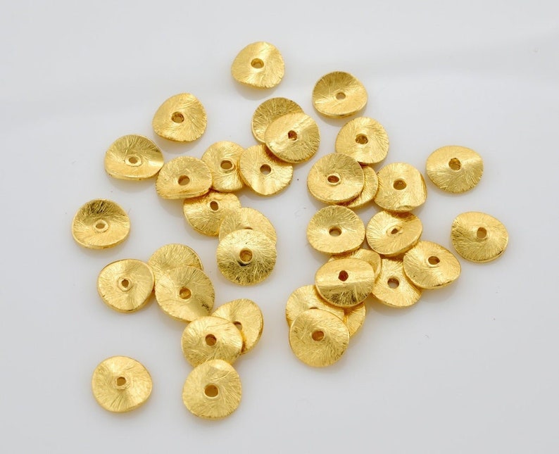 May include: A selection of small, gold-coloured, textured metal beads. Each bead is disc-shaped with a central hole, and the surface has a hammered or textured appearance. The beads are scattered on a white surface.