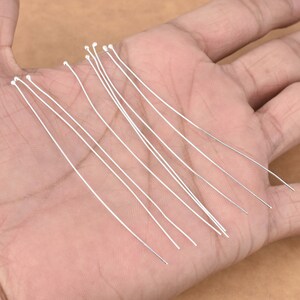 Puede incluir: Primer plano de una mano sosteniendo un manojo de alfileres de cabeza redonda finos y rectos de color plateado. Los alfileres miden aproximadamente 5 cm de largo.