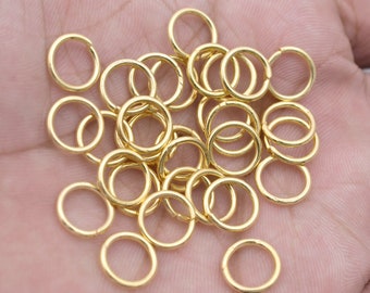 May include: A pile of gold jump rings, a type of jewelry finding, on a hand. The rings are small and round, and they are perfect for attaching charms, pendants, or other jewelry components.
