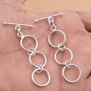Könnte beinhalten: Zwei silberne Toggle-Verschlüsse mit jeweils vier miteinander verbundenen Kreisen. Die Verschlüsse sind aus Metall und haben eine polierte Oberfläche.