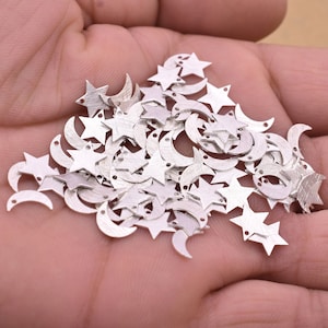 Puede incluir: Una colección de pequeños dijes metálicos plateados con forma de estrellas y medias lunas. Cada dije tiene una superficie texturizada y un pequeño agujero para ensartar. Los dijes están dispersos sobre un fondo neutro.