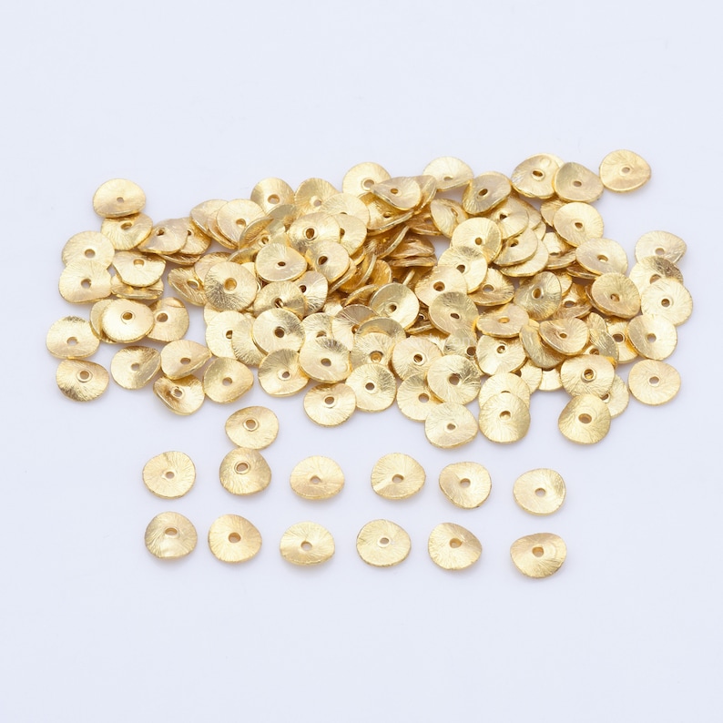 May include: A pile of small, gold-coloured, disc-shaped beads with a textured surface and a central hole. The beads are scattered on a white surface, suitable for jewellery making or other crafts.