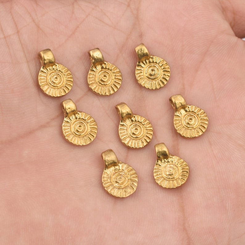 May include: Eight gold-toned metal pendant charms with a circular design and a loop at the top. The charms have a textured surface with a raised circular pattern in the centre.