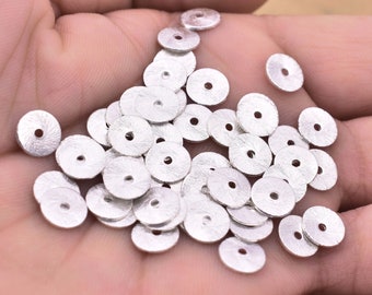 May include: A pile of silver-coloured metal beads, each with a round shape and a small hole in the centre. The beads are scattered on a hand, creating a visual representation of their size and texture.