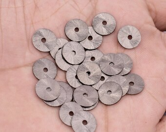 May include: A pile of silver-colored metal discs with a small hole in the center of each disc. The discs have a textured surface and are arranged in a random pattern.
