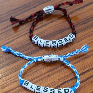 May include: Two bracelets with silver clasps and white beads spelling out the word "BLESSED". One bracelet has a black and red cord, the other has a blue and white cord.