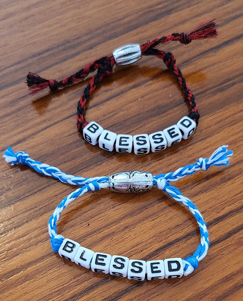 May include: Two bracelets with silver clasps and white beads spelling out the word "BLESSED". One bracelet has a black and red cord, the other has a blue and white cord.