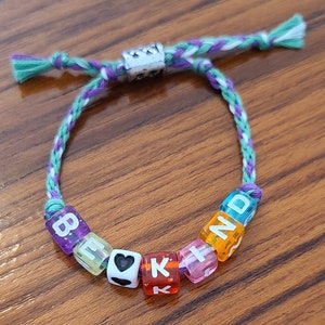 May include: A friendship bracelet with a purple, white and green braided cord and colourful letter beads spelling "BE KIND".