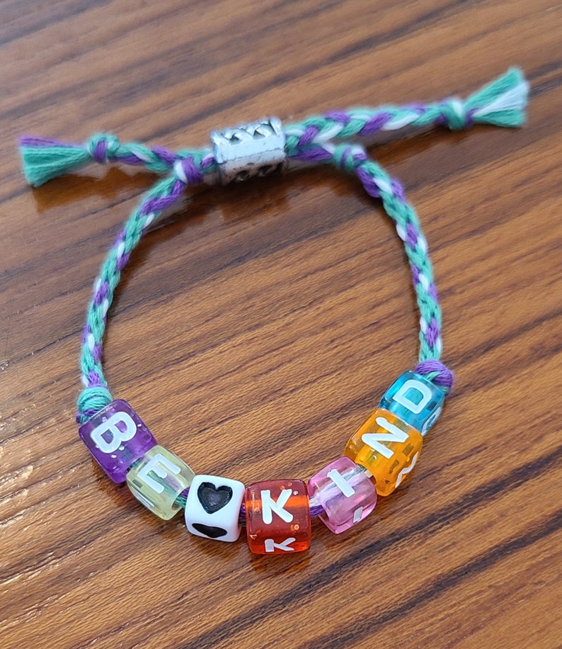 May include: A friendship bracelet with a purple, white and green braided cord and colourful letter beads spelling "BE KIND".