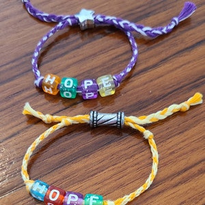 May include: Two bracelets with the word "HOPE" spelled out in colourful letter beads. The top bracelet is purple and white, while the bottom bracelet is yellow and white.