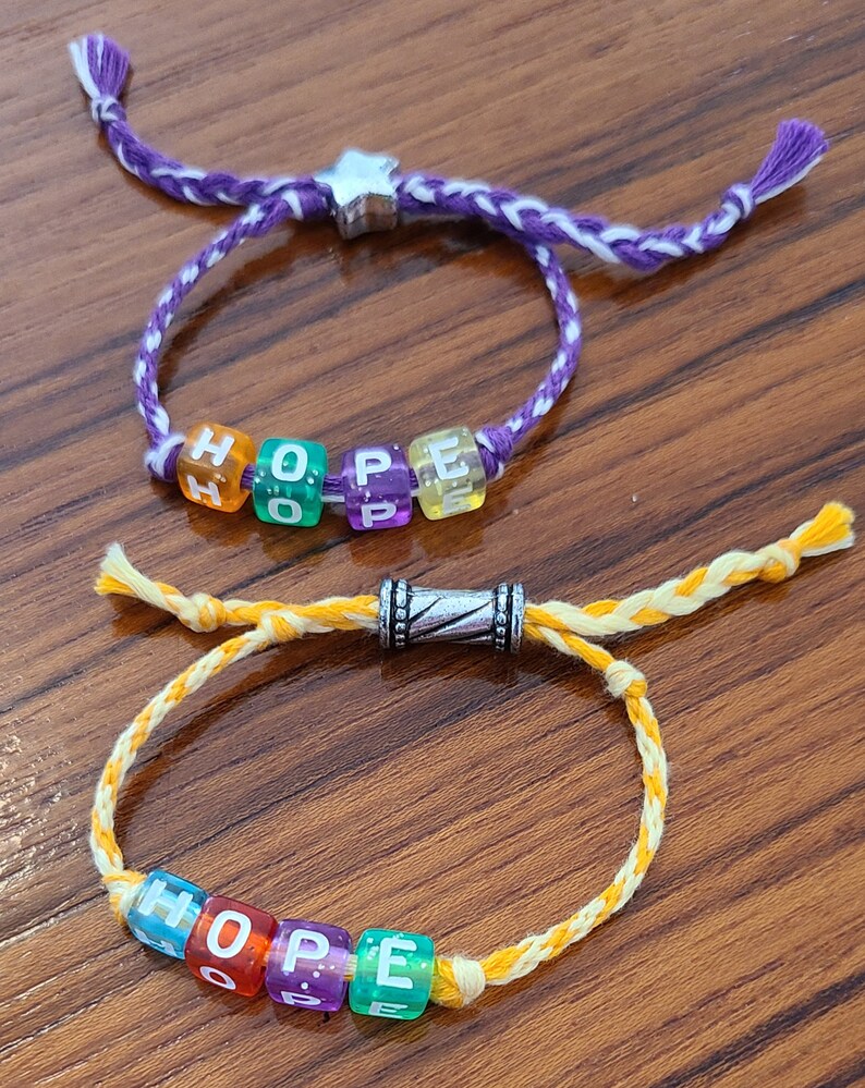 May include: Two bracelets with the word "HOPE" spelled out in colourful letter beads. The top bracelet is purple and white, while the bottom bracelet is yellow and white.