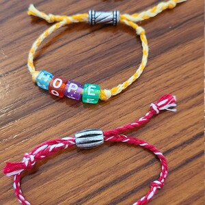 May include: Three friendship bracelets with silver bead accents. The bracelets spell out the words "FAITH", "HOPE", and "LOVE" in colourful beads.