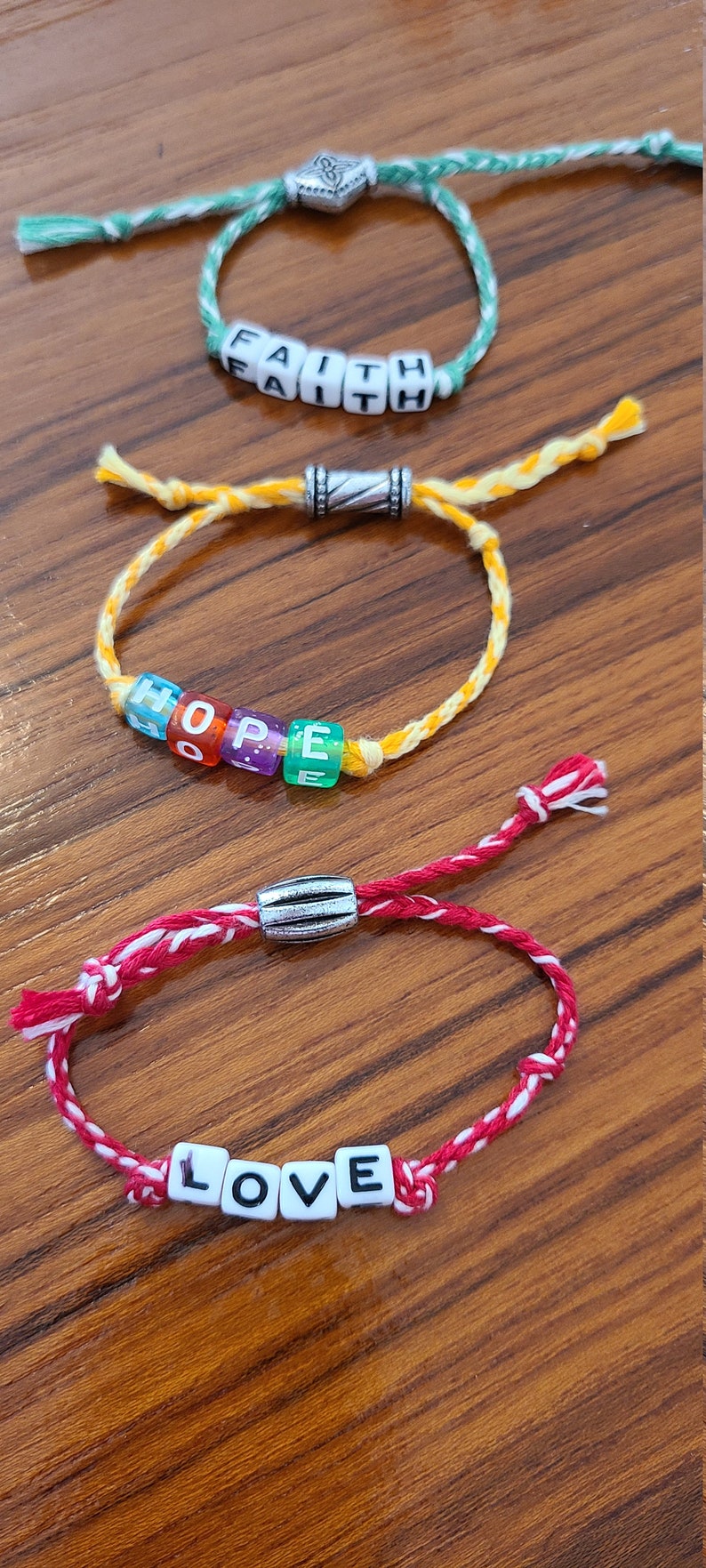 May include: Three friendship bracelets with silver bead accents. The bracelets spell out the words "FAITH", "HOPE", and "LOVE" in colourful beads.