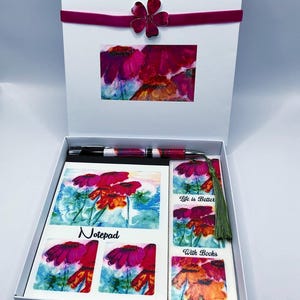 May include: A white gift box tied with a burgundy ribbon and flower clasp, containing a pen, notepad, stickers, and bookmark. The items feature a floral watercolour design in pink, red, and orange. The notepad is labelled "Notepad". The bookmark reads "Life is Better With Books".