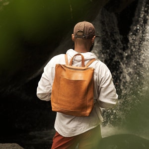 Pode incluir: Uma pessoa vestindo uma camisa branca e shorts vermelhos está perto de uma cachoeira, usando uma mochila de couro marrom.