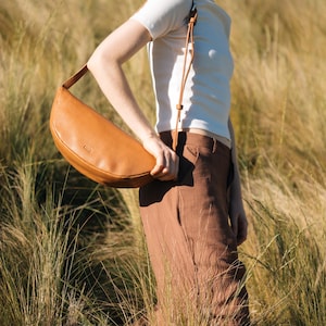 Tan full grain leather banana bag worn crossbody by woman in golden grass field, moon shape vegetable tanned sling bag handmade in Buenos Aires
