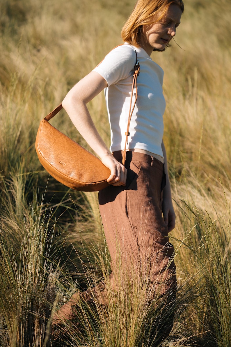 Tan full grain leather banana bag worn crossbody by woman in golden grass field, moon shape vegetable tanned sling bag handmade in Buenos Aires