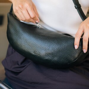 Woman seated opening black leather moon shape banana bag, full grain vegetable tanned handmade crossbody belt bag showing zip closure detail