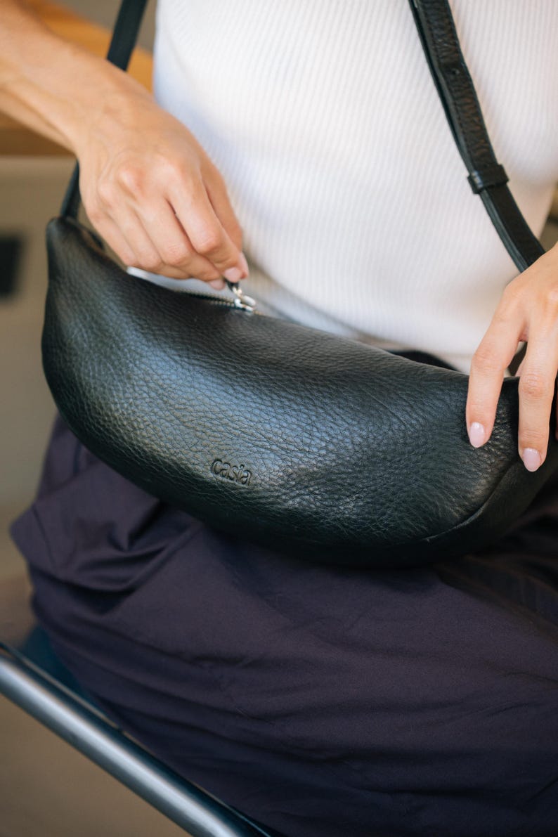Woman seated opening black leather moon shape banana bag, full grain vegetable tanned handmade crossbody belt bag showing zip closure detail