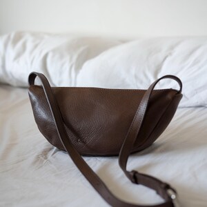 Chocolate full grain leather banana bag on white bed linen, moon shape vegetable tanned handmade crossbody sling bag lifestyle interior shot