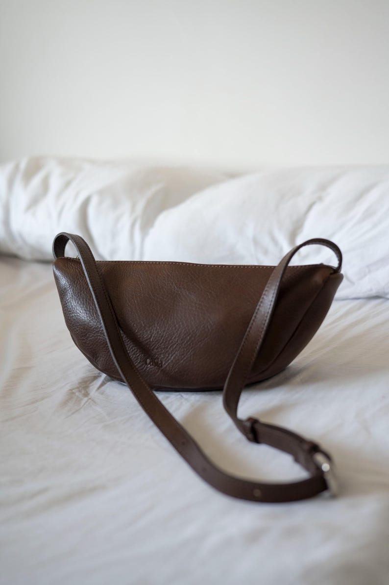 Chocolate full grain leather banana bag on white bed linen, moon shape vegetable tanned handmade crossbody sling bag lifestyle interior shot
