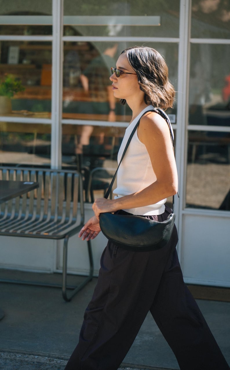Woman in urban casual outfit coat wearing black leather banana sling bag crossbody in urban setting, full grain vegetable tanned moon shape bag