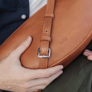 Close up of tan full grain leather banana bag showing silver buckle hardware, adjustable strap and Casia embossed logo, vegetable tanned cowhide detail