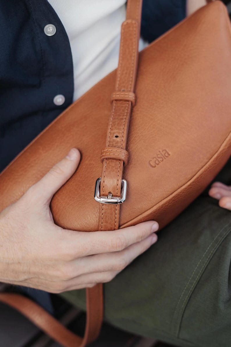 Close up of tan full grain leather banana bag showing silver buckle hardware, adjustable strap and Casia embossed logo, vegetable tanned cowhide detail