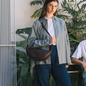 Woman in jeans wearing chocolate full grain leather banana bag crossbody, handmade vegetable tanned moon shape belt bag everyday urban lifestyle