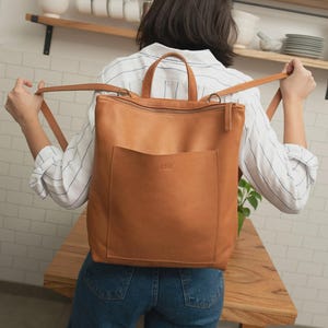 Tan full grain leather bucket-style crossbody bag with adjustable strap and aged brass hardware resting on a gray marble tabletop.