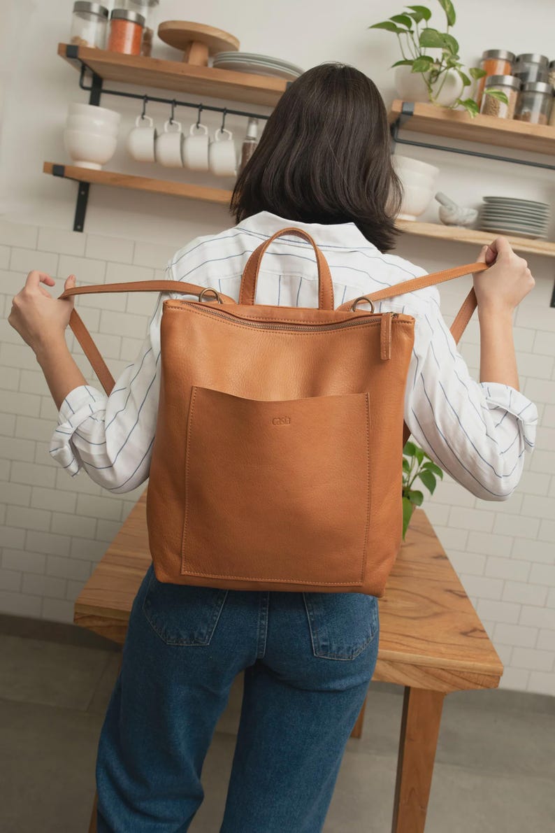 Tan full grain leather bucket-style crossbody bag with adjustable strap and aged brass hardware resting on a gray marble tabletop.