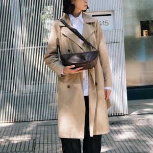 Woman in beige trench coat wearing chocolate full grain leather banana bag in urban setting, handmade vegetable tanned moon shape crossbody sling