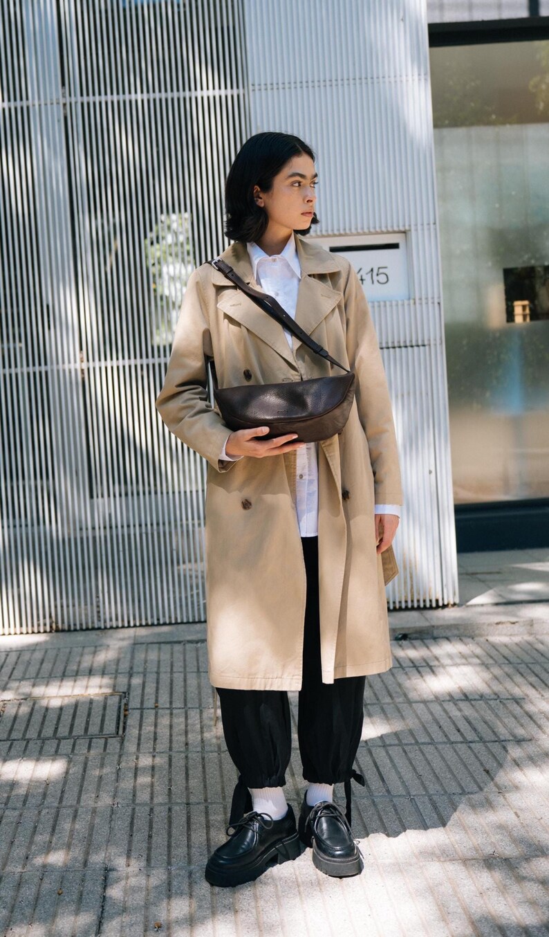 Woman in beige trench coat wearing chocolate full grain leather banana bag in urban setting, handmade vegetable tanned moon shape crossbody sling