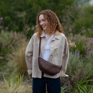 May include: A dark brown crescent-shaped crossbody bag with a smooth texture and a thin adjustable strap, worn over a beige button-down jacket, a plain white t-shirt, and dark blue denim jeans, creating a functional and layered outfit.