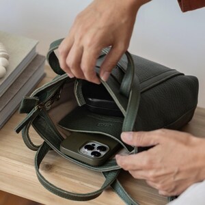 Hands opening dark leather crossbody bag on wooden table showing zipper